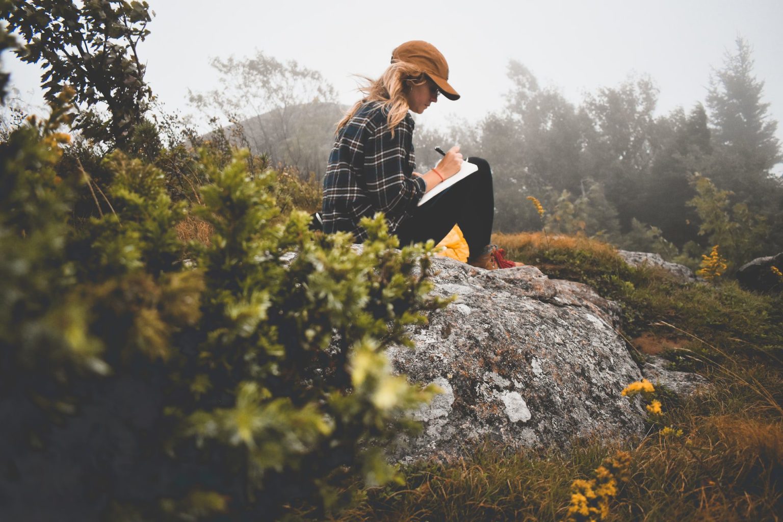 A woman outdoors sitting on a rock writing in a journal to illustrate the benefits of journaling daily in psychotherapy for borderline personality disorder