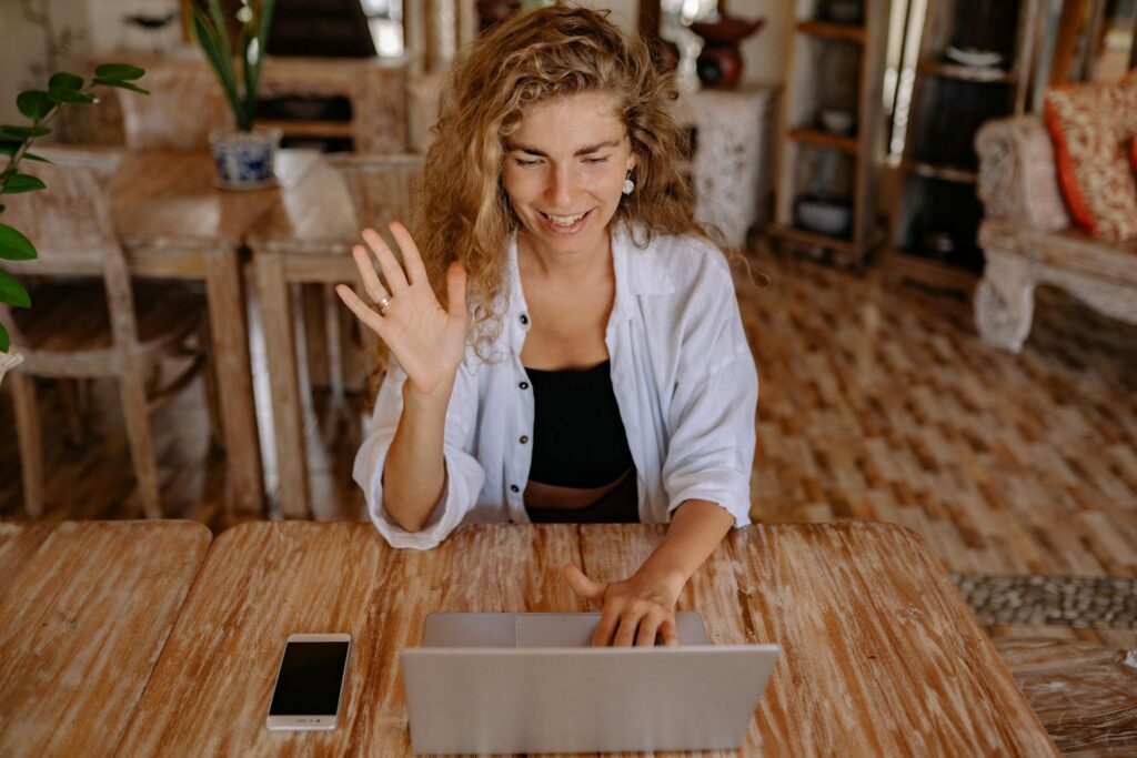 Blonde woman sitting at a table with one hand up saying hello in a video call through a laptop webcam