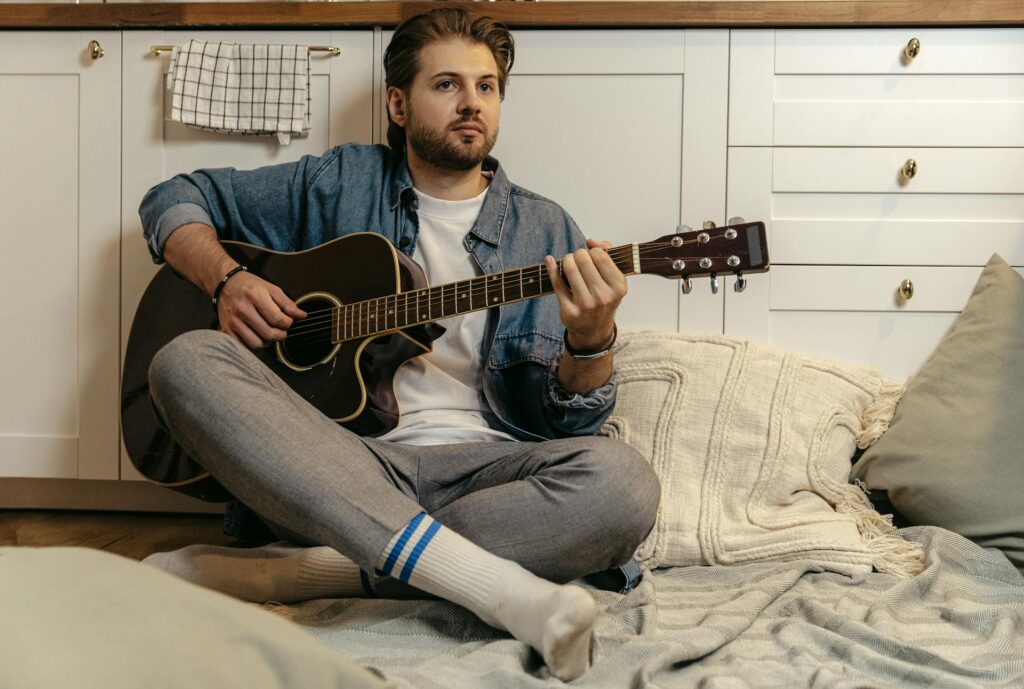 Man sitting on blankets and pillows on the floor with back against a cabinet playing a guitar