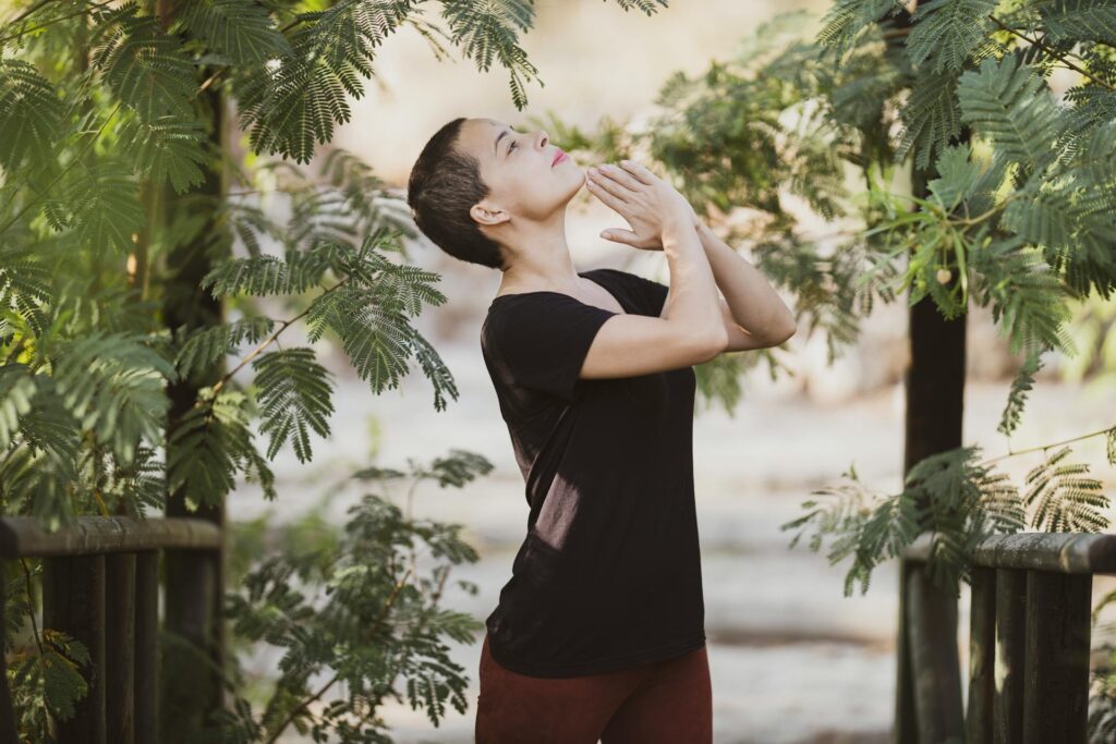 Woman standing in a leafy environment looking up at the sky with her hands together in a meditative pose