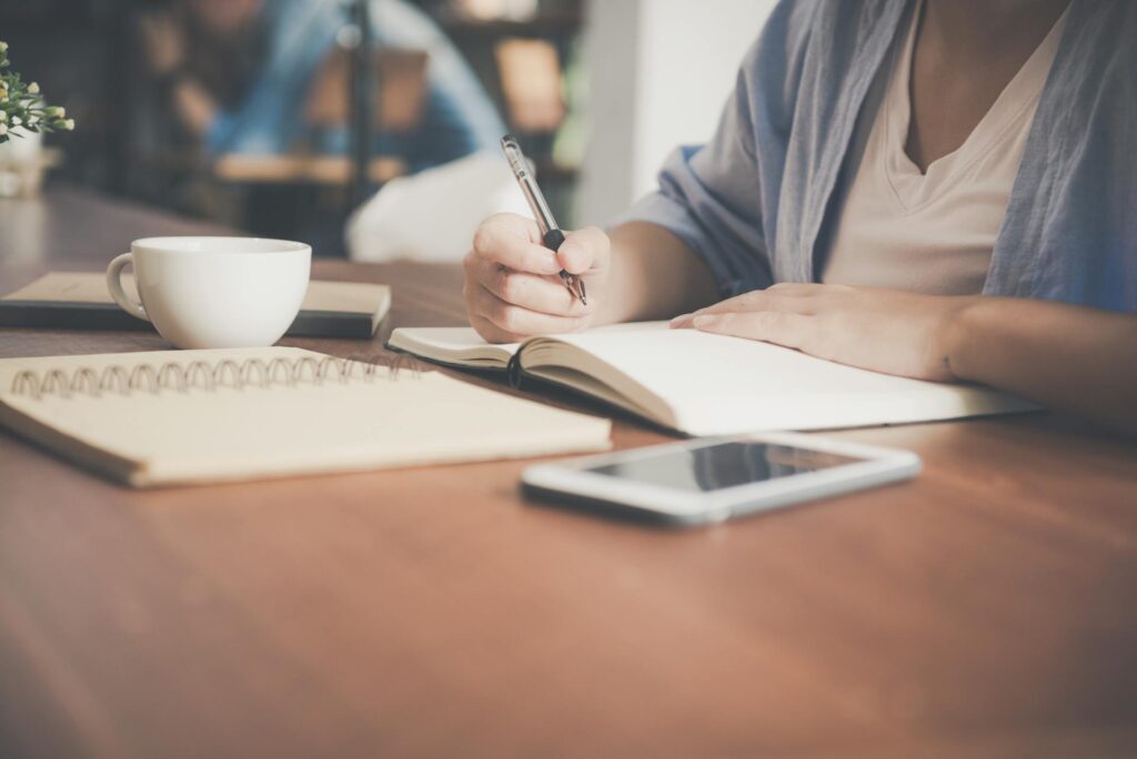 Faceless woman writing in a notebook beside a teacup and tablet